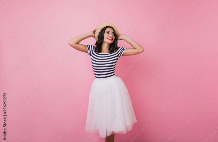 Dreamy brunette lady in long skirt expressing happiness. Lovely brunette woman dancing with smile and touching her hat.